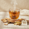 A double-walled clear glass cup filled with amber tea and a bloomed flower, set on a wooden coaster with dried tea ingredients and a hanging tag reading 'Buds Bloom' on a light wood surface.