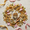 A styled flat-lay of a glass jar filled with loose herbal tea, surrounded by scattered dried flowers, chamomile, pink petals, and a small wooden scoop on a light fabric background with ribbon accents.