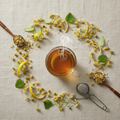 A steaming glass cup of amber tea arranged on a textured beige fabric, surrounded by loose-leaf chamomile flowers, lemon slices, green leaves, two wooden spoons holding loose tea, and a metal tea infuser.