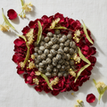 A top-down flat lay of small oval brownish-green seeds arranged in a circle on a white fabric background, surrounded by deep red flower petals and small pale yellow blossoms.