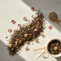 Loose-leaf herbal tea blend labeled 'Blissful Bloom' scattered on a white marble surface, mixed with dried pink rose petals and small yellow flowers. A small bowl of the tea blend, a brass mesh strainer, and a cream ribbon tag are arranged nearby.